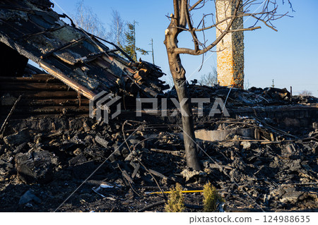 Charred remains of a building lie in a forest area, revealing destruction caused by a recent wildfire, with blackened trees and rubble visible Charred remains of a building lie in a forest area, revealing destruction caused by a recent wildfire, with blackened trees and rubble visible 124988635