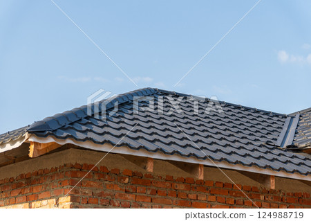 Workers diligently install black tile roofing on a brick house under a vibrant blue sky, showcasing craftsmanship and sunny weather 124988719