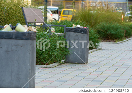 Two trash bins overflow with litter beside a bench in a city park, surrounded by green plants during daylight hours Two trash bins overflow with litter beside a bench in a city park, surrounded by green plants during daylight hours 124988730