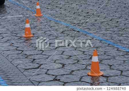 Orange traffic cones sit on a wet cobblestone surface, marking no parking zones and creating safety measures in rainy conditions Orange traffic cones sit on a wet cobblestone surface, marking no parking zones and creating safety measures in rainy conditions 124988740