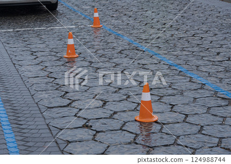 Wet pavement in an urban parking lot features orange cones spaced evenly, indicating reserved spaces during a rainy day 124988744