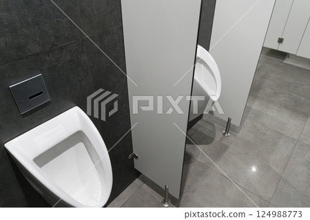 A contemporary restroom showcases a white urinal beside closed privacy stalls, all set against dark gray walls and tiled flooring 124988773