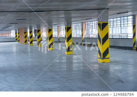 An expansive parking garage features yellow and black safety barriers on the columns, illuminated by natural light from large windows 124988784