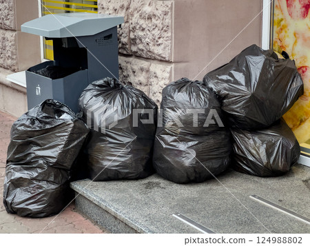 Multiple black trash bags are piled on the sidewalk next to a litter bin in a city setting Multiple black trash bags are piled on the sidewalk next to a litter bin in a city setting 124988802