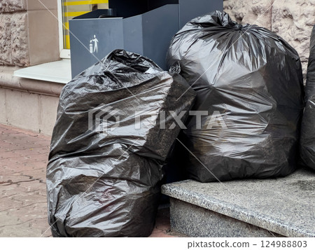 Several black trash bags sit next to a waste bin on a busy urban sidewalk, showing signs of recent litter collection efforts Several black trash bags sit next to a waste bin on a busy urban sidewalk, showing signs of recent litter collection efforts 124988803