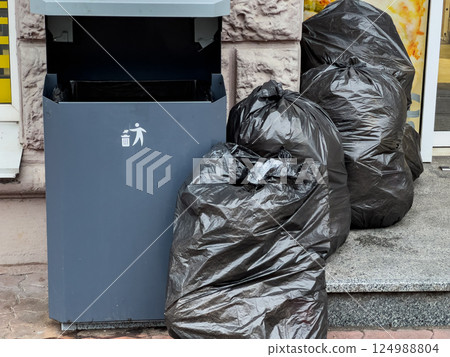 Black trash bags are stacked beside a public waste bin near a store entrance, highlighting urban waste management issues during the day Black trash bags are stacked beside a public waste bin near a store entrance, highlighting urban waste management issues during the day 124988804