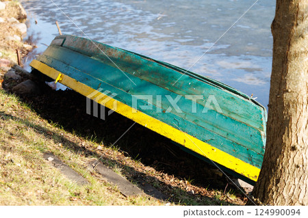 Old green and yellow boat turned upside down on lakeside grass near tree trunk. Early spring outdoor photography. Rustic fishing concept. Design for postcard, wallpaper, greeting card, banner 124990094