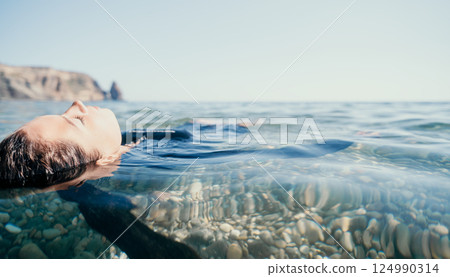 Man Floating Ocean Water Relaxation - A man relaxes in the clear blue ocean water with a rocky shoreline in the distance. Man Floating Ocean Water Relaxation - A man relaxes in the clear blue ocean water with a rocky shoreline in the distance. 124990314
