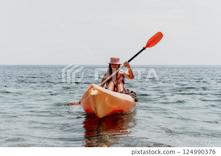 Kayaking Woman Paddle Ocean - A woman wearing a pink hat kayaks on the ocean, paddling with an orange paddle. Kayaking Woman Paddle Ocean - A woman wearing a pink hat kayaks on the ocean, paddling with an orange paddle. 124990376