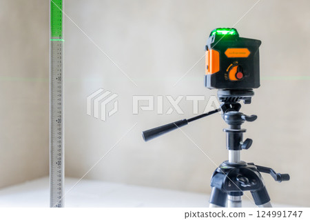 Laser level on tripod projecting green beam onto ruler in empty room Laser level on tripod projecting green beam onto ruler in empty room 124991747