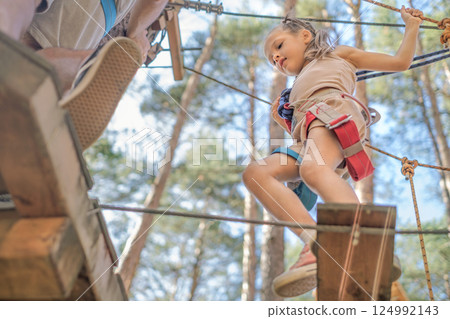 A brave little girl climbs a rope track from a suspension bridge in an Entertainment park 124992143