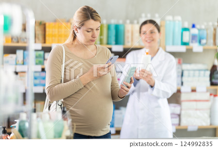 Pregnant woman in pharmacy scans QR code on antifungal cream, pharmacist offer similar product in background. 124992835