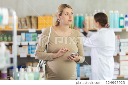 Pregnant woman checking prescription details on phone in pharmacy Pregnant woman checking prescription details on phone in pharmacy 124992923