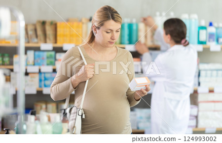 Pregnant woman choose pills of supplements in pharmacy, staff work in background Pregnant woman choose pills of supplements in pharmacy, staff work in background 124993002
