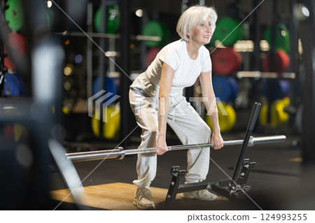 Senior woman performing deadlifts with barbell at gym 124993255