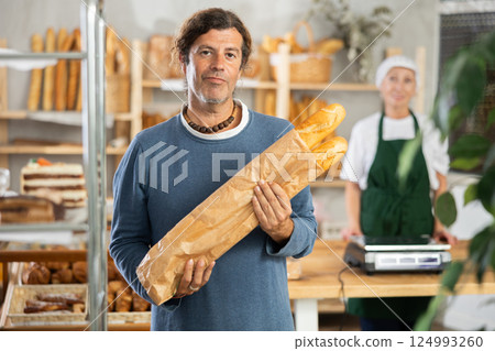 Man buyer with baguettes in package stands near showcase in bakery Man buyer with baguettes in package stands near showcase in bakery 124993260