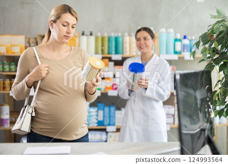 In drug store, pregnant woman examines formula for feeding, pharmacist offer item in background In drug store, pregnant woman examines formula for feeding, pharmacist offer item in background 124993564