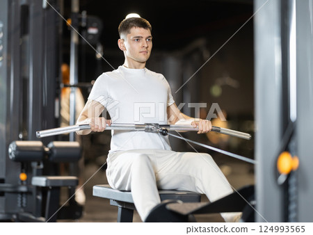 In gym, young man uses cable row machine to strengthen arm muscles 124993565