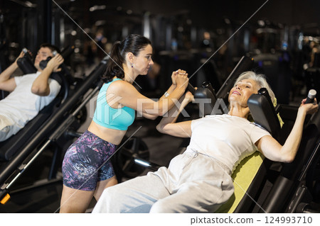 Senior woman performing squats on hack machine with coach 124993710