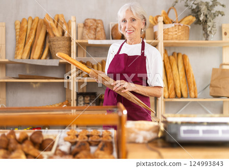 Mature saleswoman holding paper bag with baguettes in bakery 124993843