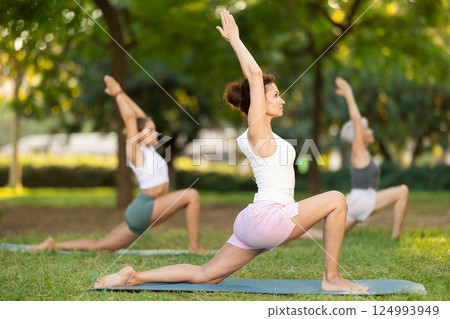 Skilled happy young female teacher demonstrating variation of warrior pose of yoga for group women at city park Skilled happy young female teacher demonstrating variation of warrior pose of yoga for group women at city park 124993949