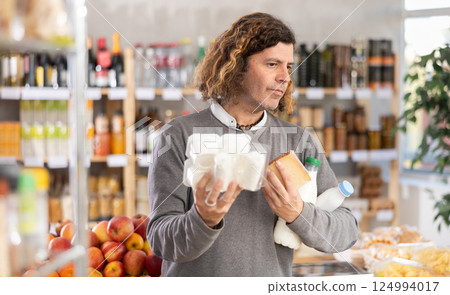 Male shopper with various dairy products in her hands continues to make choice in grocery department of supermarket Male shopper with various dairy products in her hands continues to make choice in grocery department of supermarket 124994017