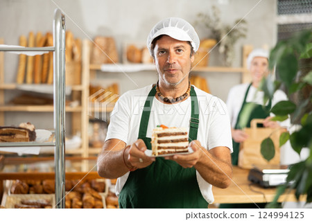 In bakery, man seller holds piece of puff pastry cake on disposable plate. 124994125