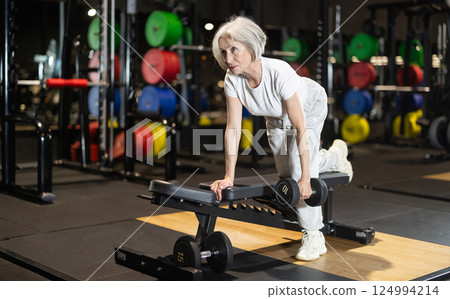 Senior woman performing one-arm dumbbell row on bench 124994214