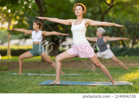 Young woman with group of active women exercising full body stretching workout during yoga classes in green park 124994238