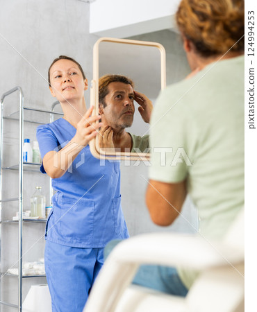 Man scrutinizing his own reflection in mirror held by female cosmetologist Man scrutinizing his own reflection in mirror held by female cosmetologist 124994253