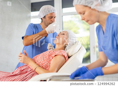 Male doctor with help of nurse performs rejuvenation procedure for elderly female in forehead area 124994262