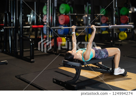 Woman fitness club client lying on bench, lifts barbell Woman fitness club client lying on bench, lifts barbell 124994274
