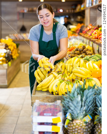 Female seller holding bananas standing in fruit and vegetable section of supermarket Female seller holding bananas standing in fruit and vegetable section of supermarket 124994327