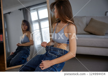 Anorexic slim body woman wearing big jeans standing front of mirror 124994434