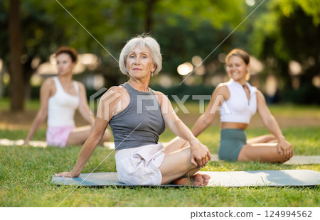 Sedulous old lady and group of women doing spinal twist pose of yoga on mat in light city park together Sedulous old lady and group of women doing spinal twist pose of yoga on mat in light city park together 124994562
