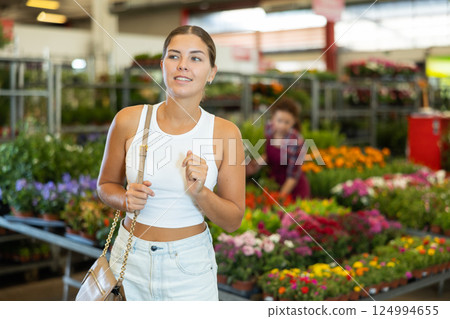 Customer girl stands in aisle between rows with variety of flowering plants in pots Customer girl stands in aisle between rows with variety of flowering plants in pots 124994655