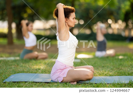 Portrait of caucasian woman sitting on sports mat and making yoga meditation in lotus pose and hands up with group together in park at daytime 124994667