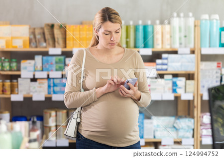 Young woman scrolling phone while looking for something in drugstore 124994752