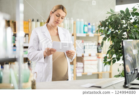 Pregnant woman employee in white coat works in pharmacy sales area using computer. 124994880
