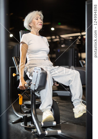 In gym, senior woman uses gym apparatus to strengthen leg muscles In gym, senior woman uses gym apparatus to strengthen leg muscles 124994898