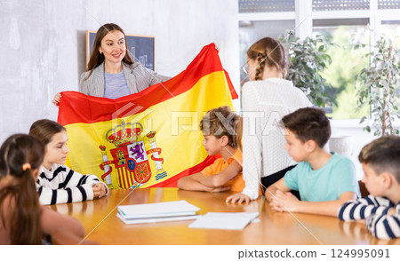 Excited female teacher discussing with preteens students state flag of Spain in auditory of middle school Excited female teacher discussing with preteens students state flag of Spain in auditory of middle school 124995091
