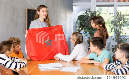 Group of curious preteen learners and young female teacher with flag of Morocco in classroom of academy Group of curious preteen learners and young female teacher with flag of Morocco in classroom of academy 124995092