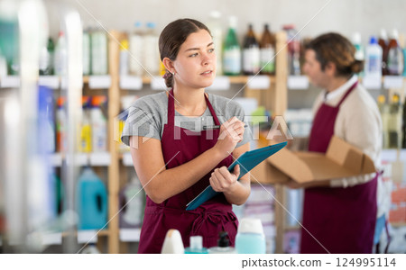 Female manager makes notes on an paper - conducts an inventory. In background, male salesperson places goods on shelves 124995114