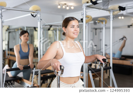 Young positive woman stretches her body muscles while sitting on a special chair in Pilates gym 124995393