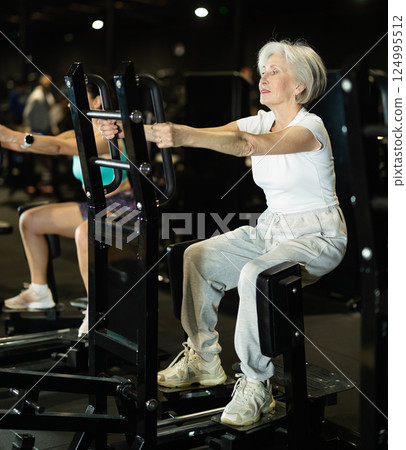 Elderly woman using standing abductor machine during workout at gym 124995512