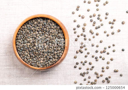 Puy lentils in a wooden bowl on linen. Dried variety of small lentils, mottled, of slate-gray to dark green color, with blue marbling. Lens esculenta variety, grown in the Auvergne region of France. 125000654