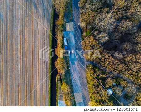 Aerial view of a quiet road cutting through farmland and forest, with a parked car and cyclists nearby. Contrasting textures of plowed field and dense trees highlight the rural landscape. Aerial view of a quiet road cutting through farmland and forest, with a parked car and cyclists nearby. Contrasting textures of plowed field and dense trees highlight the rural landscape. 125001571