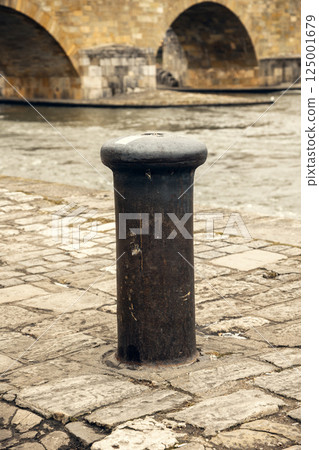A worn mooring bollard on a historic cobblestone riverbank, set against the backdrop of an ancient 125001679