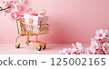 Golden shopping cart with a gift box and pink blossoms on a soft pink background with copy space 125002165