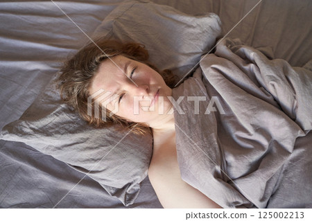Caucasian female adult relaxing in bed with cozy grey bedding and natural light 125002213